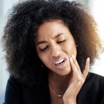 Curly-haired woman in a black shirt cringes in pain and touches her cheek due to a toothache dental emergency