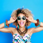 Black woman in summer clothes smiles while wearing sunglasses against a blue wall