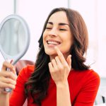 Brunette woman in a red blouse smiles at herself in a handheld mirror after teeth whitening
