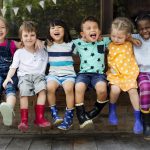 A group of young children sit on a bench together and smile with their arms around each other